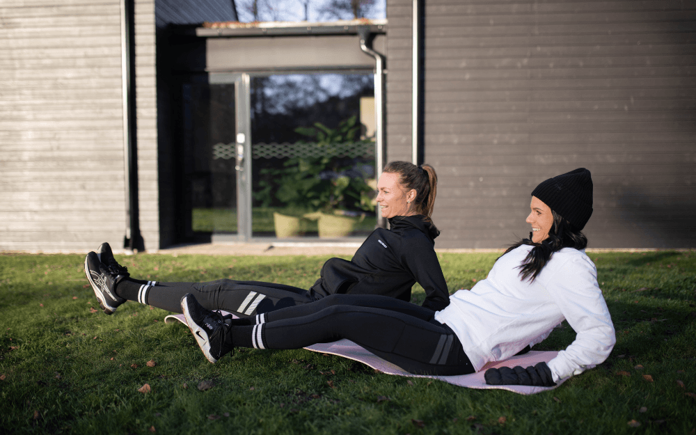 Två kvinnor som gör benglid på träningsmatta utomhus.