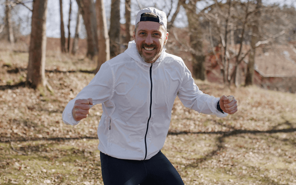 Löpcoach Micke står leende i skogen med vit tröja, bakptvänd keps och knytna nävar framför sig.