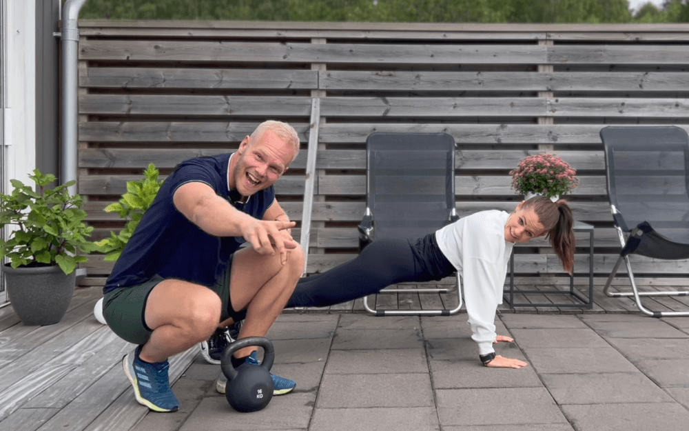 Kvinna gör plankan medan PT sitter på huk bredvid med Kettle Bells framför sig.