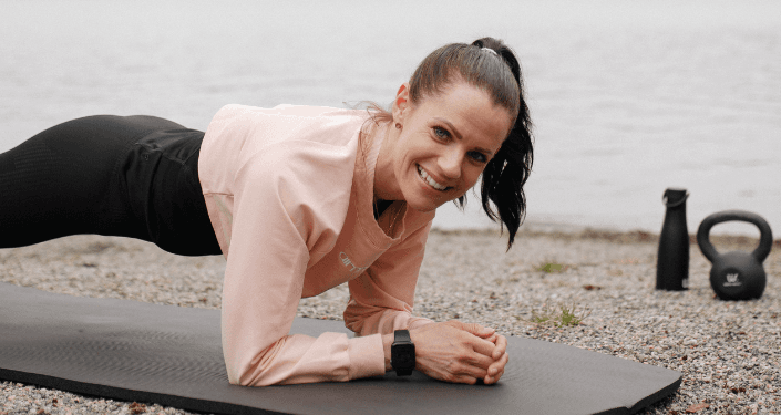 Leende kvinna med rosa tröja gör plankan på en strand.