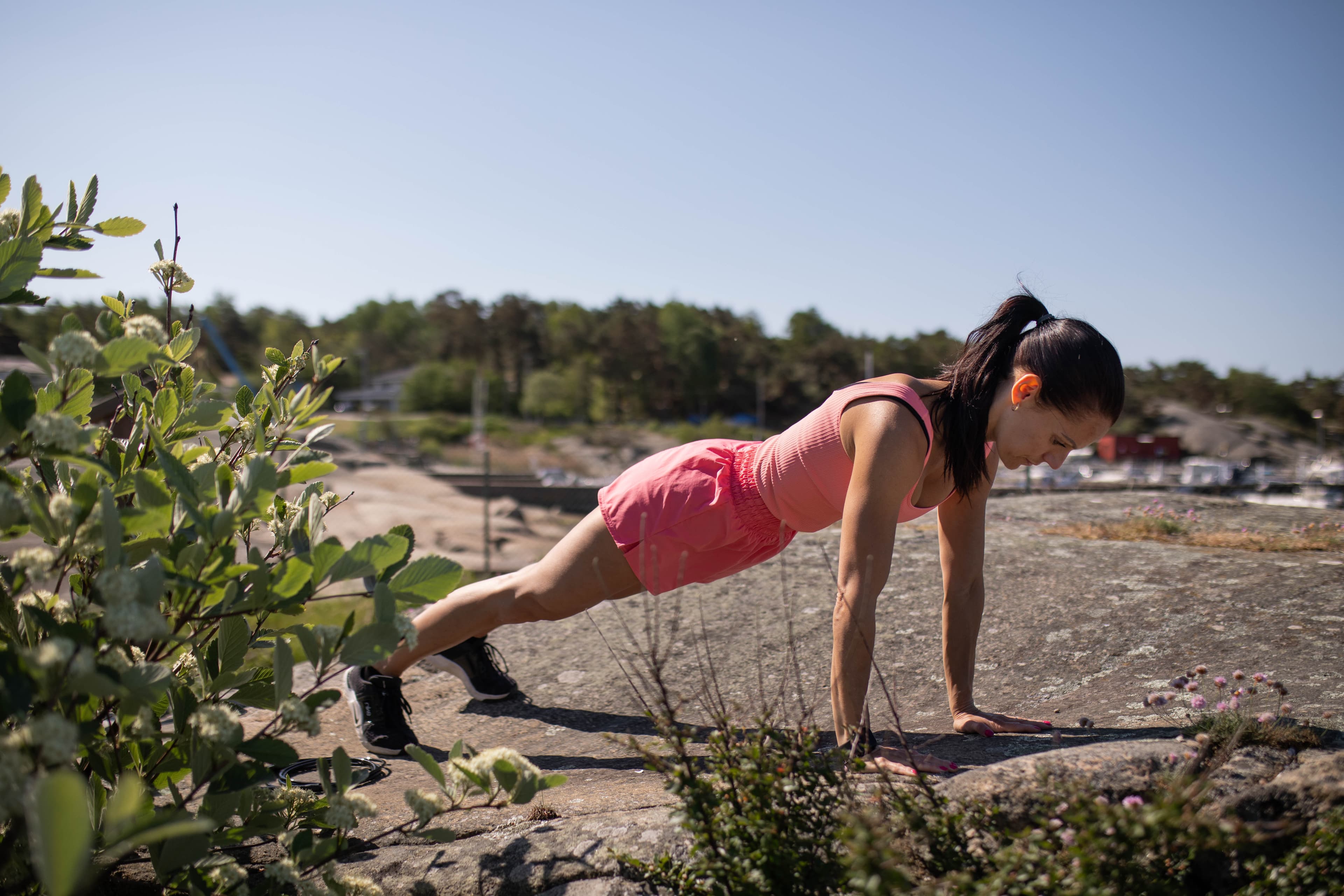 Kvinna i rosa shorts och rosa linne gör armhävningar på en klippa i vackert väder.