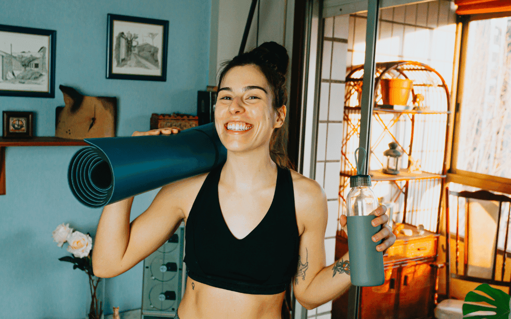 Leende, mörkhårig kvinna står i hemmet med träningsmatta på axeln och vattenflaska i handen.