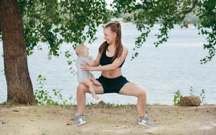 En kvinna gör sumo-benböj med en bebis på sitt knä. I bakgrunden syns havet och ett träd.