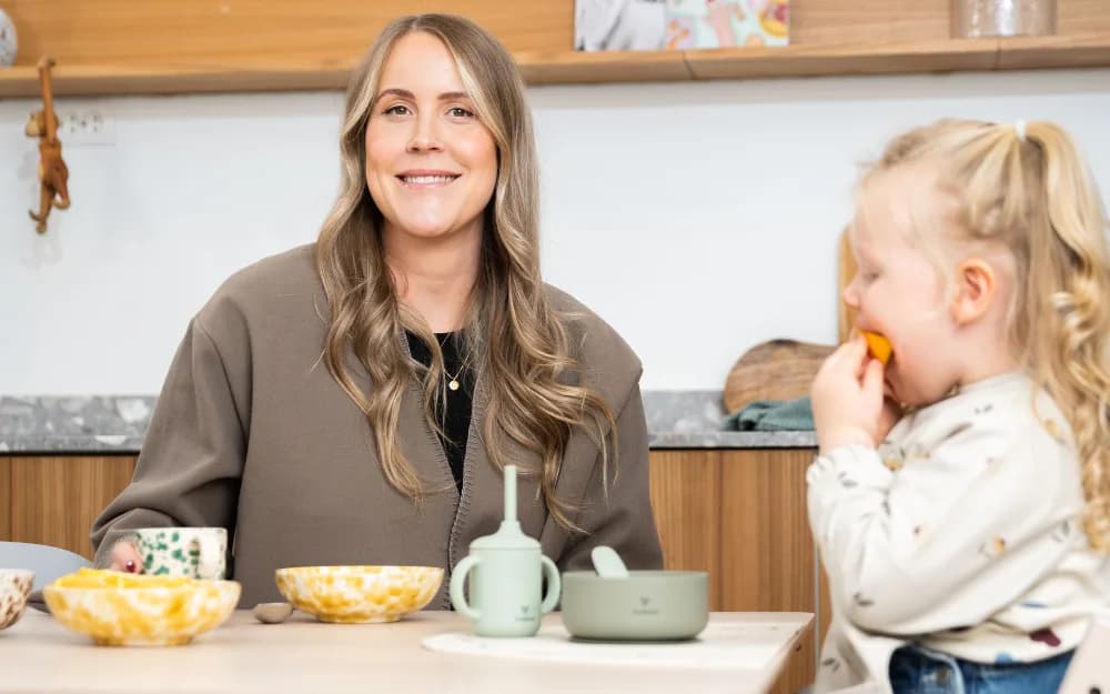 Elin Oresten som driver företaget Knatteplock sitter vid köksbordet och ler mot kameran. Hennes dotter sitter bredvid och äter plockmat.