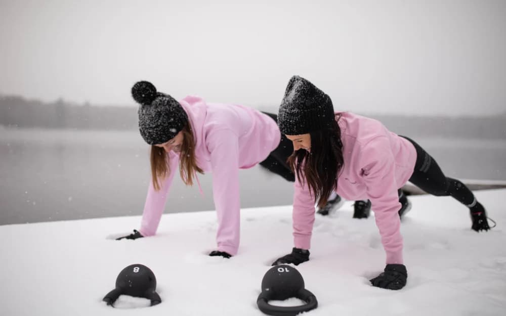 Två kvinnor i rosa tröjor och svart mössa gör armhävningar i snön.