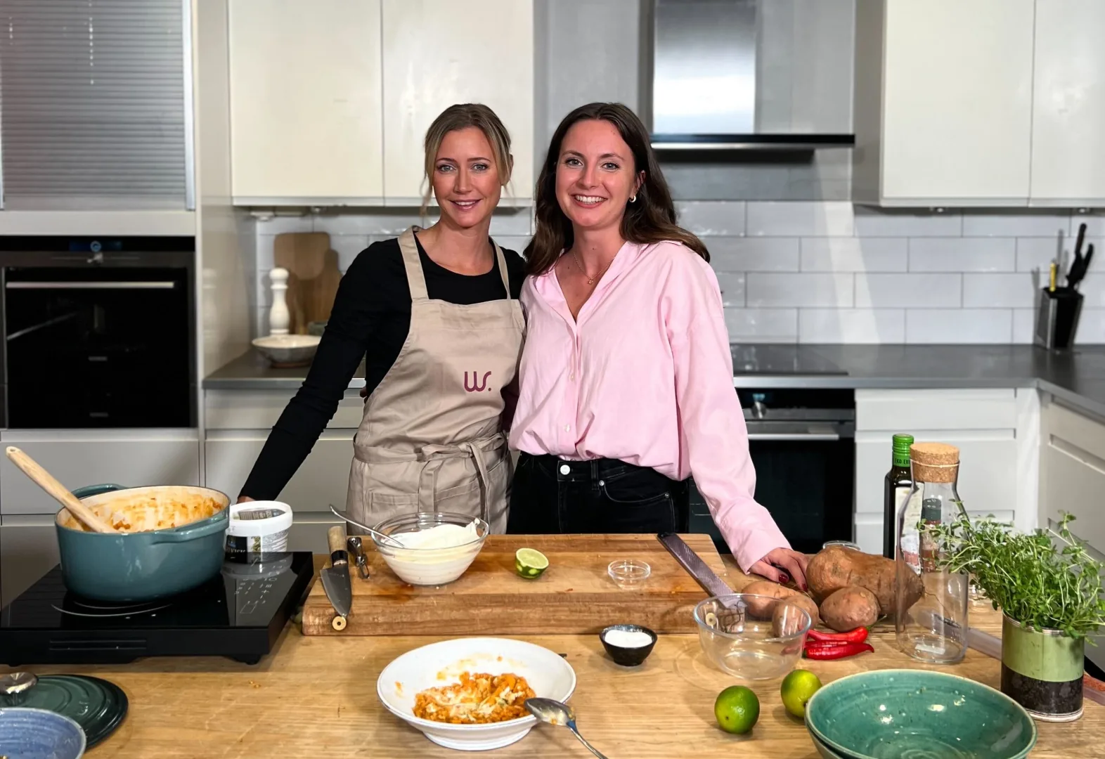 Catarina König och Joanna Ostrowska lagar mat i ett kök. På stekplattan står en gryta och det finns andra råvaror på bordet.