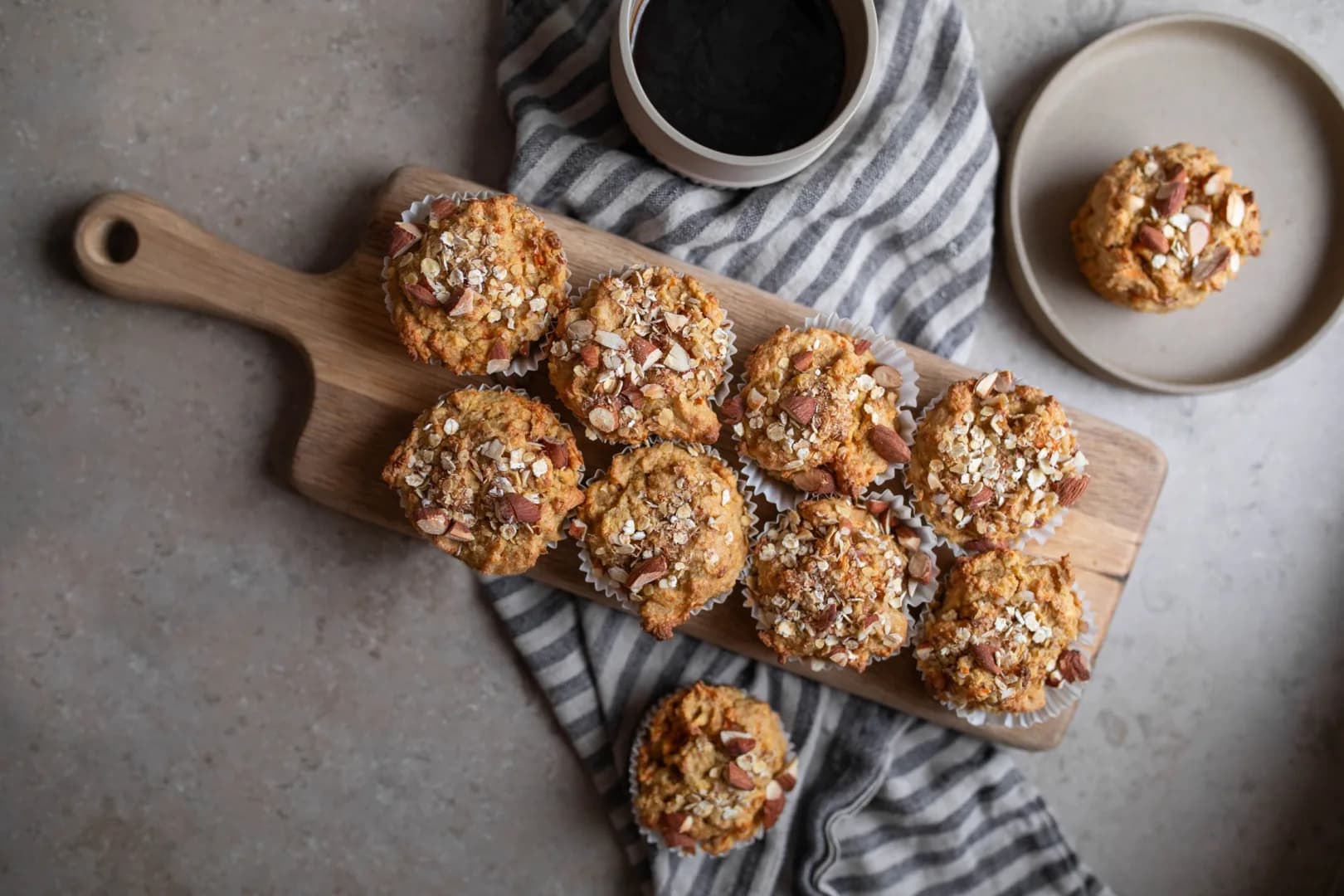 Gräddade muffins med nötter på en skärbräda i trä. I bakgrunden syns en kopp kaffe och en svartvitrandig kökshanduk.
