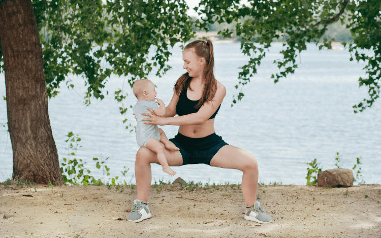 En kvinna gör sumo-benböj med en bebis på sitt knä. I bakgrunden syns havet och ett träd.